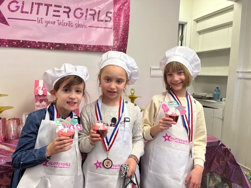 Three girls in chef hats holding cupcakes under the Glitter Girls banner